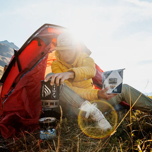 MX3 Pasta Carbonara retkiruokapussi keltatakkisen naisen kädessä, joka istuu maassa punaisen teltan edessä ja lämmittää vettä retkikeittimellä auringon paisteessa.