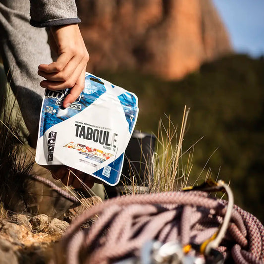 Itämainen salaatti Taboule retkiruokapussi kädessä, takana aurinkoinen vuorenseinämä ja edessä kiipeilyköysiä.