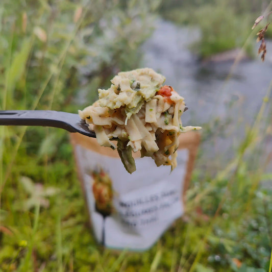 Pad Thai-nuudelit kasviksilla retkiruokaa valmiina haarukassa, takana retkiruokapussi ja joki.