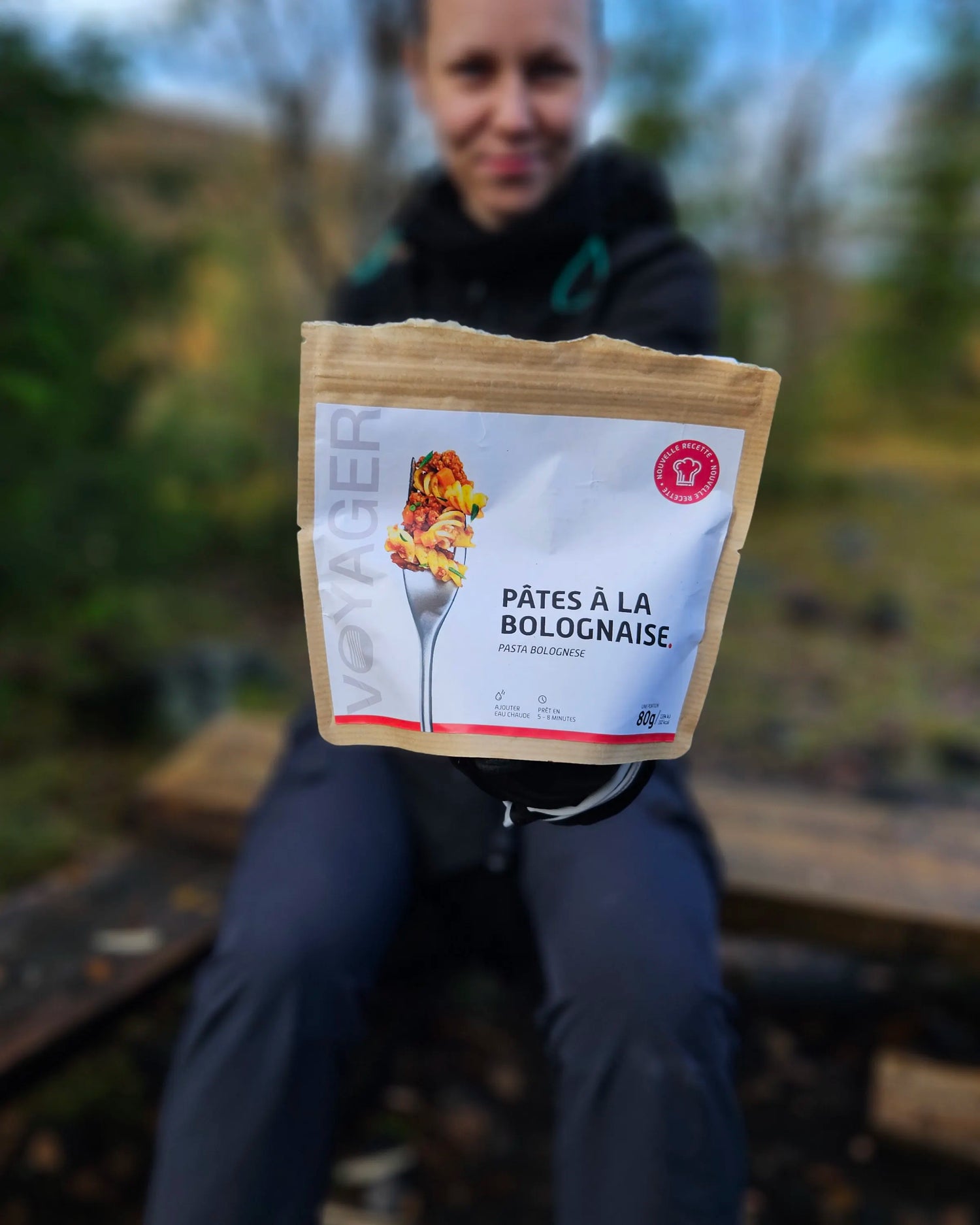 Nainen istumassa puupenkillä, ojentaa kohti kameraa Pasta Bolognese retkiruokapussia
