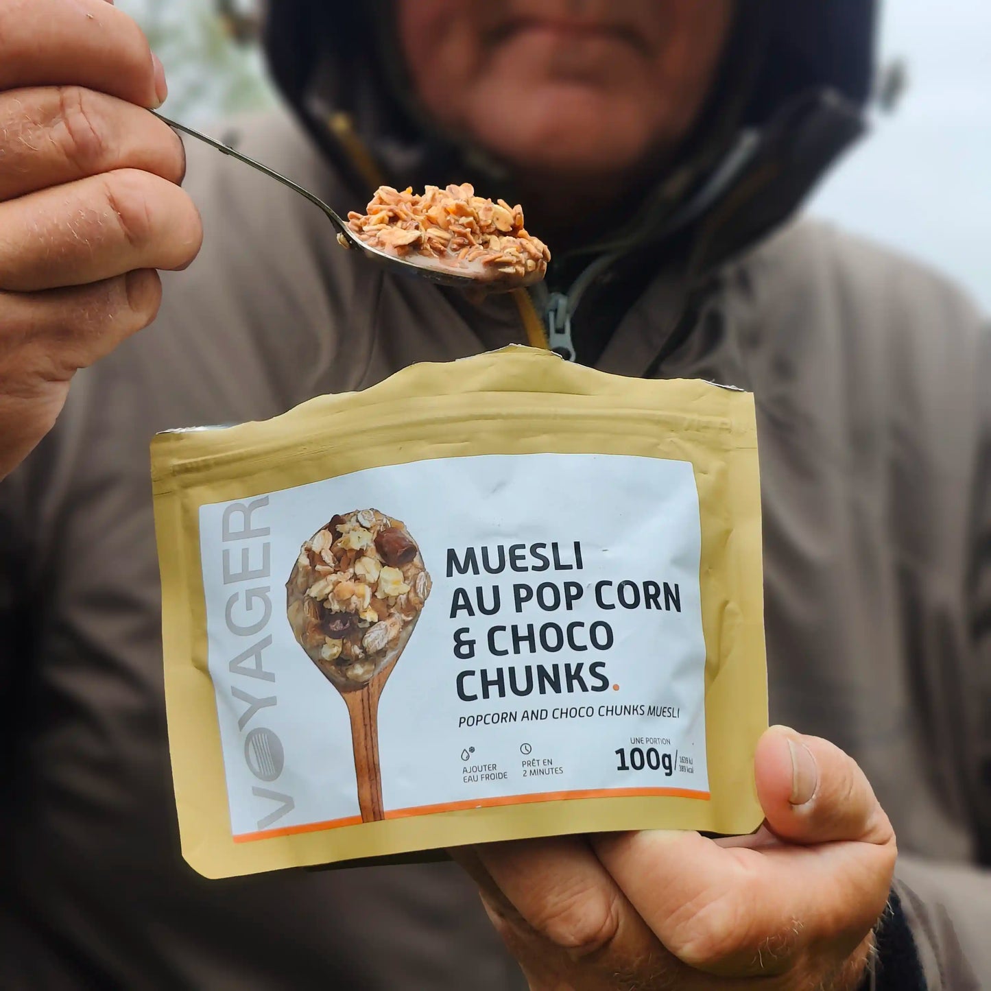 Suklaa-popcornmysli retkiruoka-aamupalapussi ja lusikallinen Suklaa-popcornmysli-retkiruokaa valmiina lusikassa ruskeatakkisen miehen kädessä.