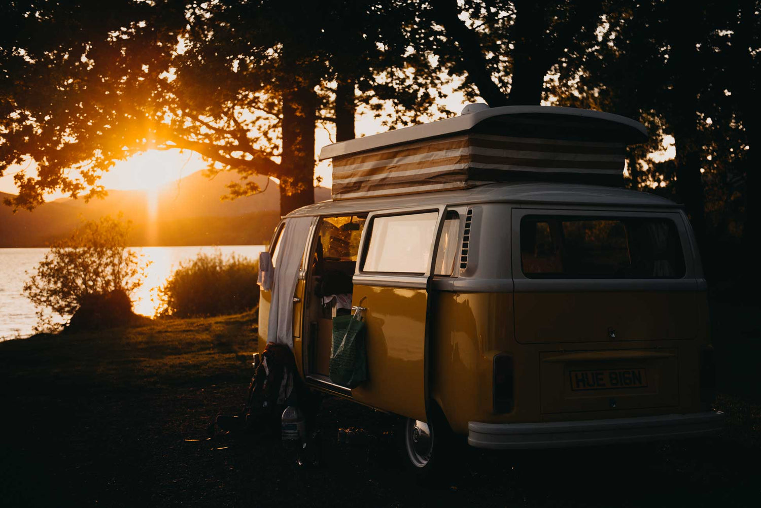 Oranssi-valkoinen Wolksvagen Camper auto järven rannalla auringonlaskussa.