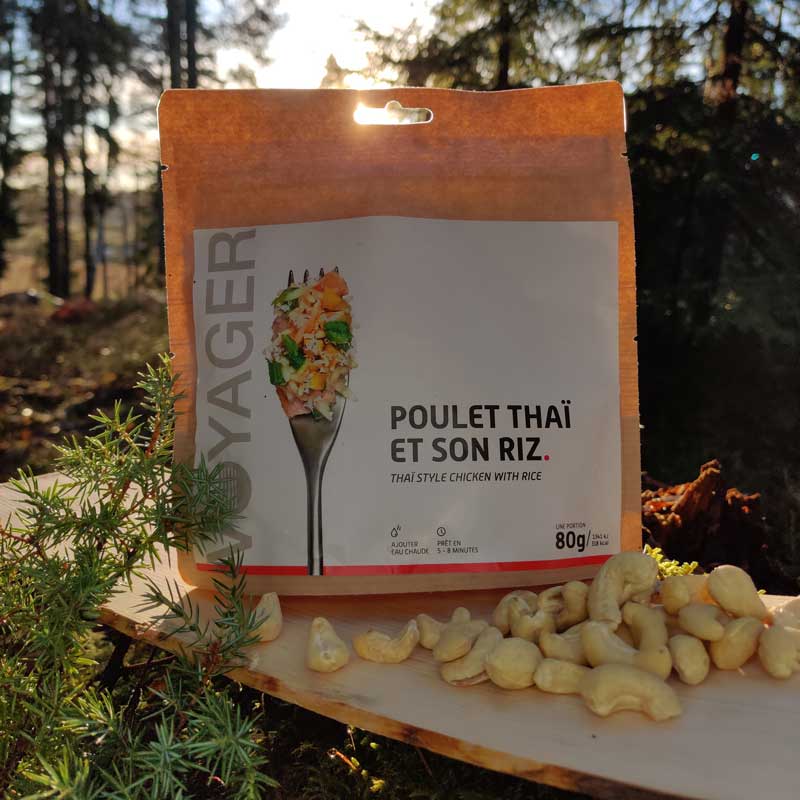 Hedelmäinen kana riisillä retkiruokapussi havujen keskellä päreen päällä vastavalossa, edessä cashew-pähkinöitä.