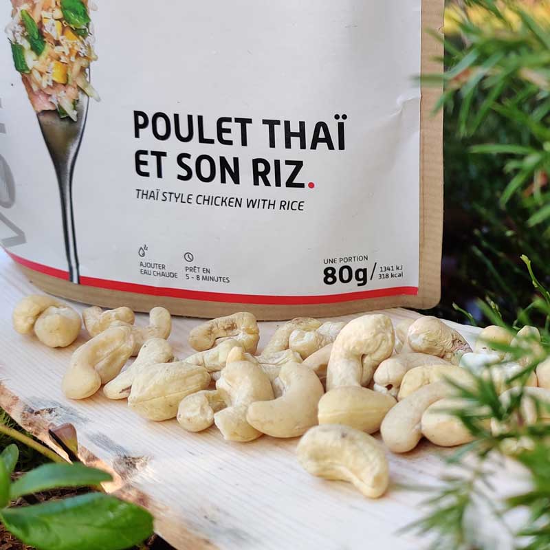 Hedelmäinen kana riisillä retkiruokapussi havujen keskellä päreen päällä, edessä cashew-pähkinöitä.
