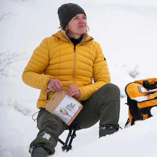Juustoinen kinkku-perunasoselaatikko retkiruoka kierrätettävässä pussissa keltatakkisen ja vihreä housuisen vaalean naisen kädessä, joka istuu lumisessa ympäristössä.