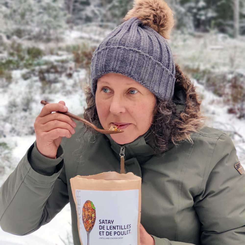 Nainen vihreässä takissa ja harmaassa pipossa syö kana-linssisatay retkiruokaa talvisessa maisemassa.