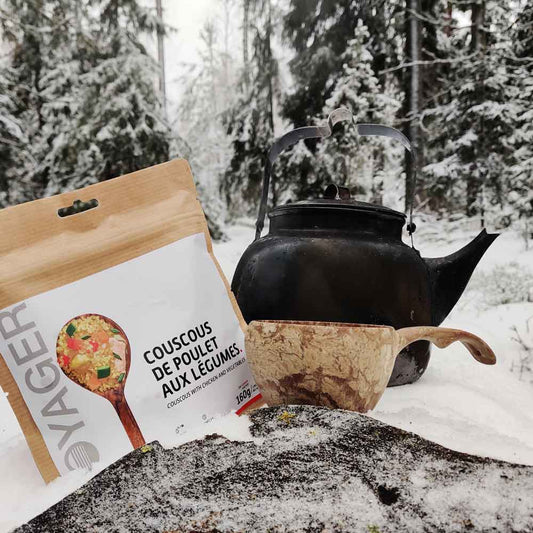 Kana-couscous ja vihanneksia-retkiruokapussi lumisessa maassa, vieressä kupilka-kuksa ja nokipannu.