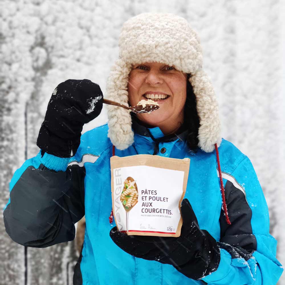 Nainen sinisessä takissa ja karvalakissa syömässä kanapastaa kesäkurpitsalla-retkiruokaa lummisateessa.