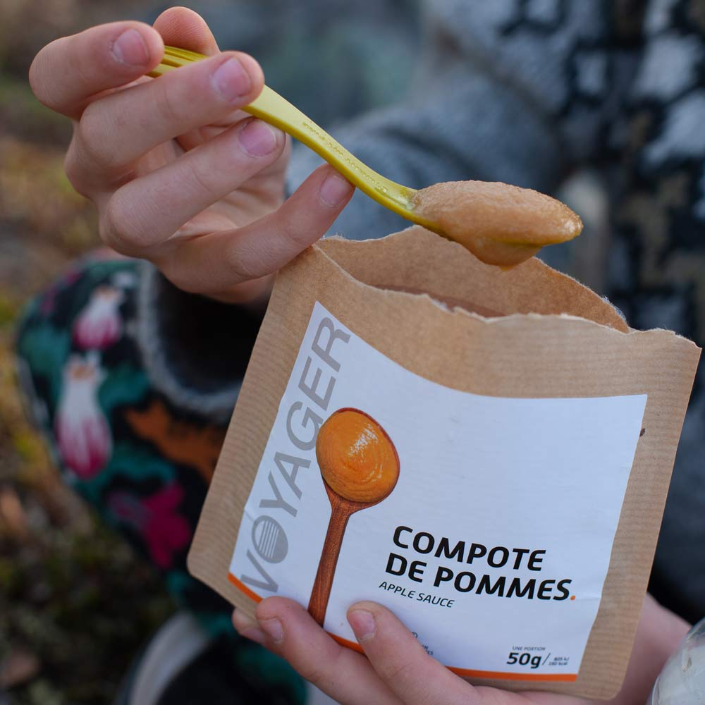 Omenakompotti retkiruokapussi kädessä ja omenakompottia keltaisessa lusikassa.