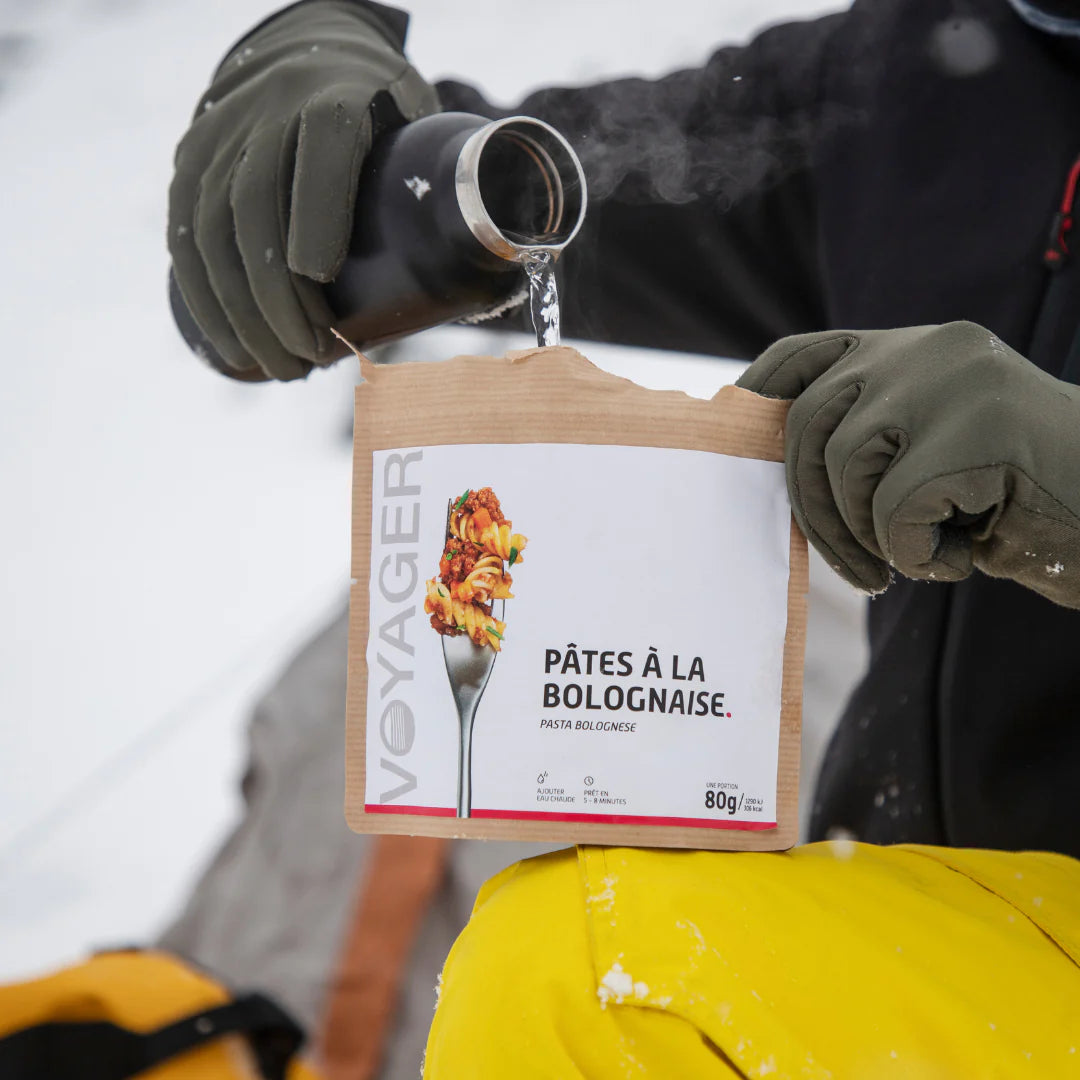 keltahousuinen ja mustatakkinen ihminen kaataa kuumaa vettä termospullosta pasta bolognese retkiruokaan kierrätettävässä pussissa talvella.