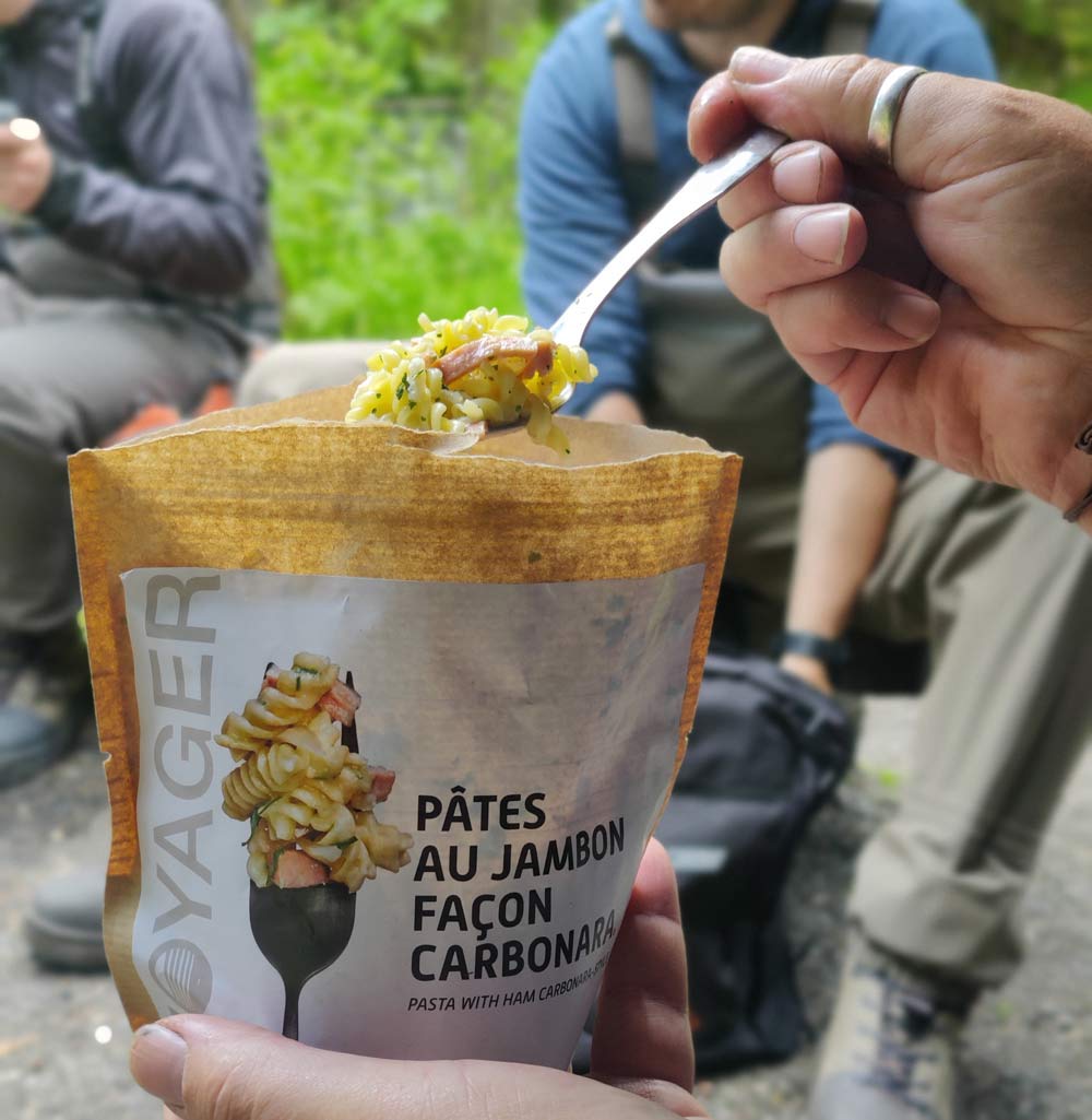 Pasta carbonara retkiruoka lusikassa leirinuotion äärellä, taustalla ihmisiä istumassa.