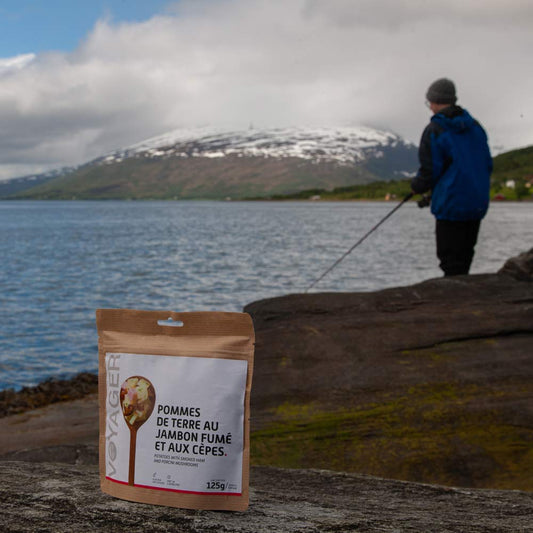 Perunapataa savukinkulla ja tateilla-retkiruoka pussissa edessä, taustalla kalastaja, meri ja lumihuippuinen tunturi.