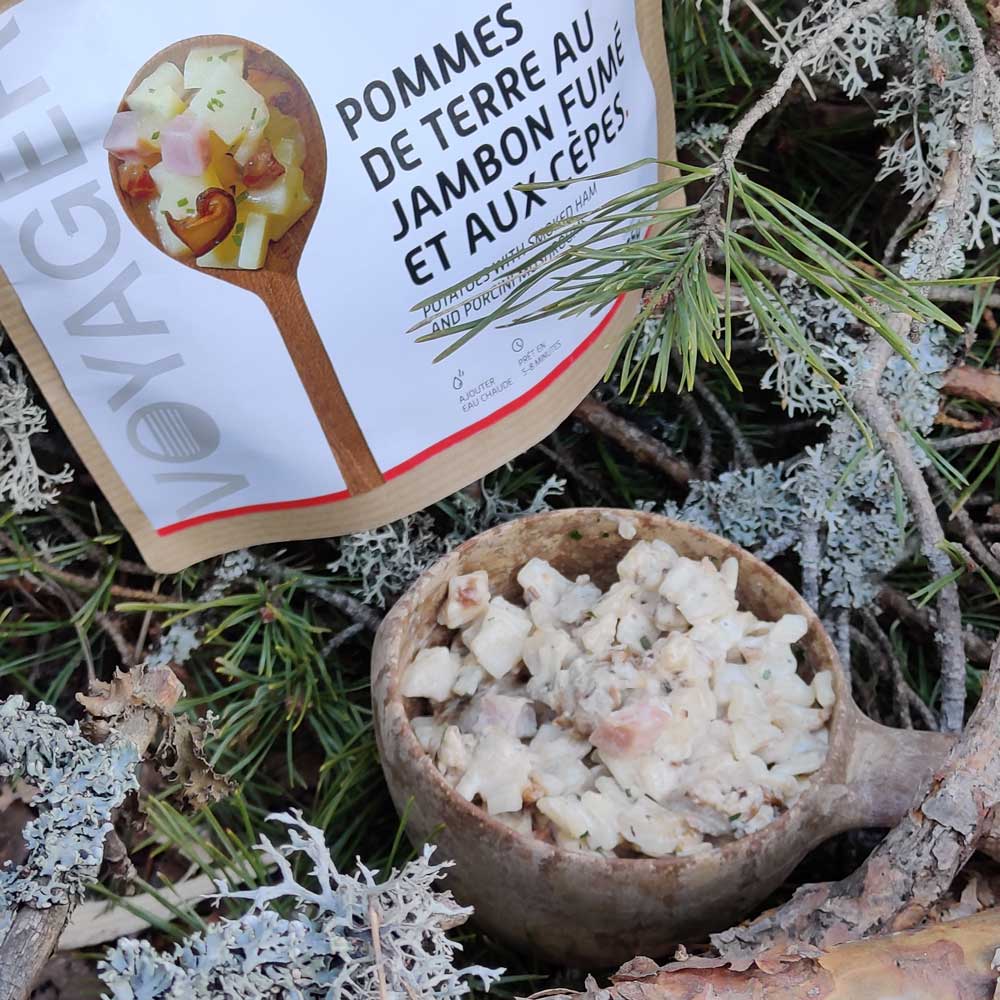 Perunapataa savukinkulla ja tateilla Kupilka-astiassa oksien ja havujen keskellä, takana retkiruokapussi.