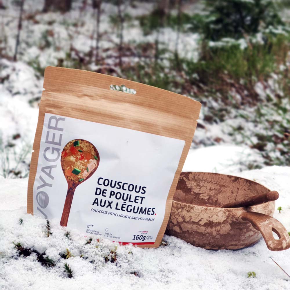 Kana-couscous ja vihanneksia-retkiruokapussi lumisessa maassa, vieressä kupilka-astia.