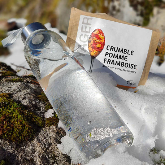 Omena-vadelma jälkiruokapaistos, retkiruoka kierrätettävässä pussissa lumisella kalliolla, edessä lasinen vesipullo.