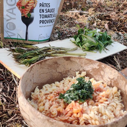 Pasta Provencal retkiruoka valmiina Kupikla-astiassa, taustalla retkiruokapussi ja villivihanneksia päreen päällä.