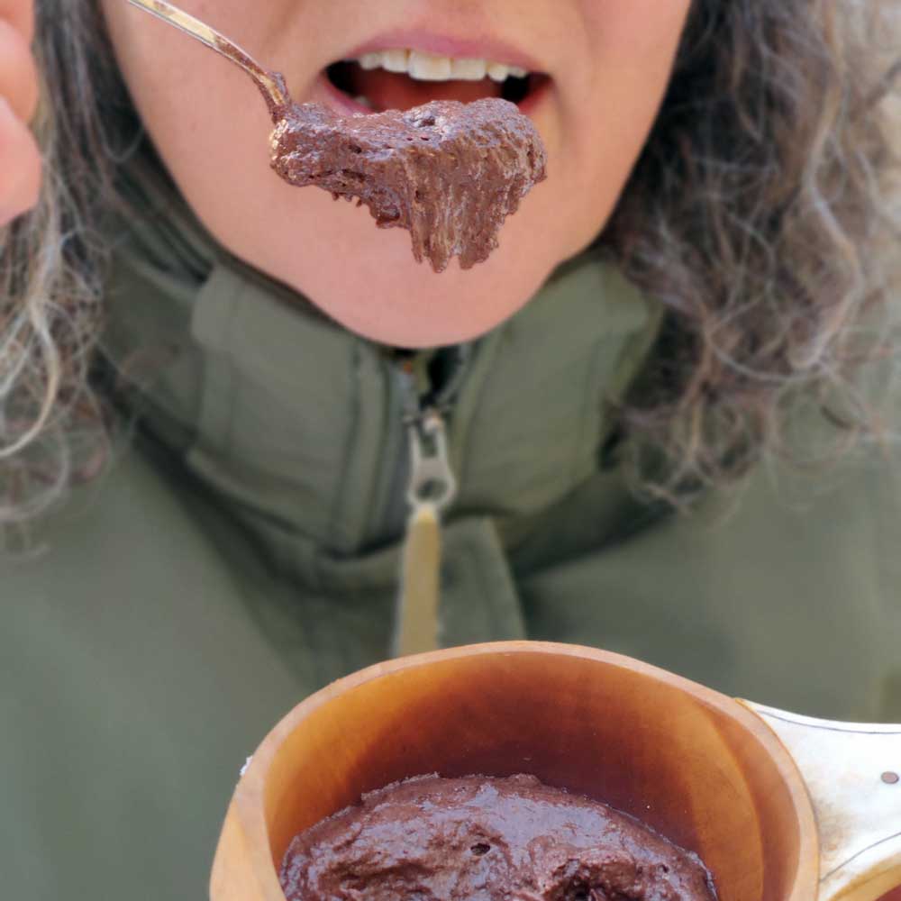 Vihreä takkinen nainen syömässä suklaamousse suklaahipuilla-retkiruoka puisesta kuksasta lusikalla.