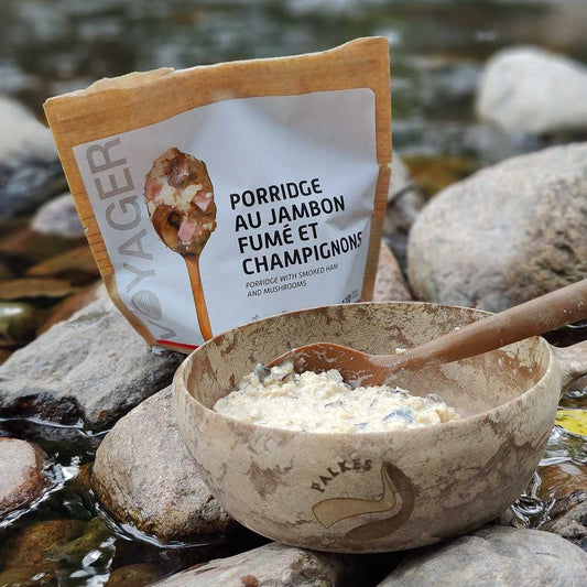 Vaeltajan savukinkku-herkkusienipuuro-retkiruoka valmiina Kuplikassa, jossa lusikka kivien päällä veden ääressä., takana retkiruokapussi.