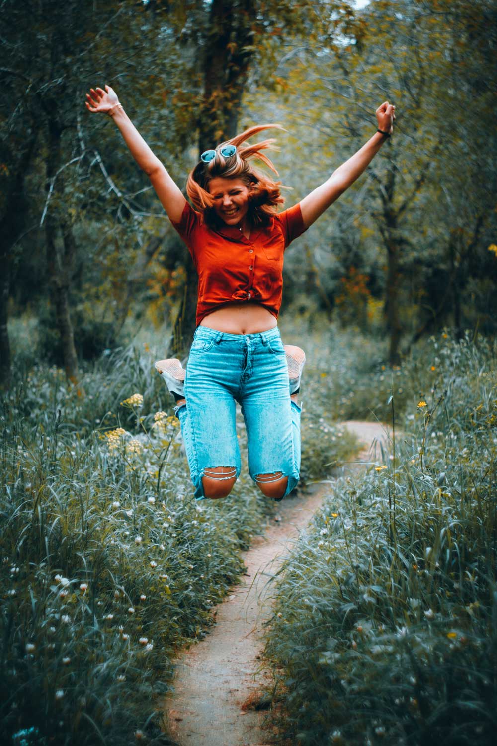 Nainen farkuissa ja punaisessa t-paidassa hyppää ilmaan innoissaan metsäpolulla ja ojentaa käsiään kohti taivasta.