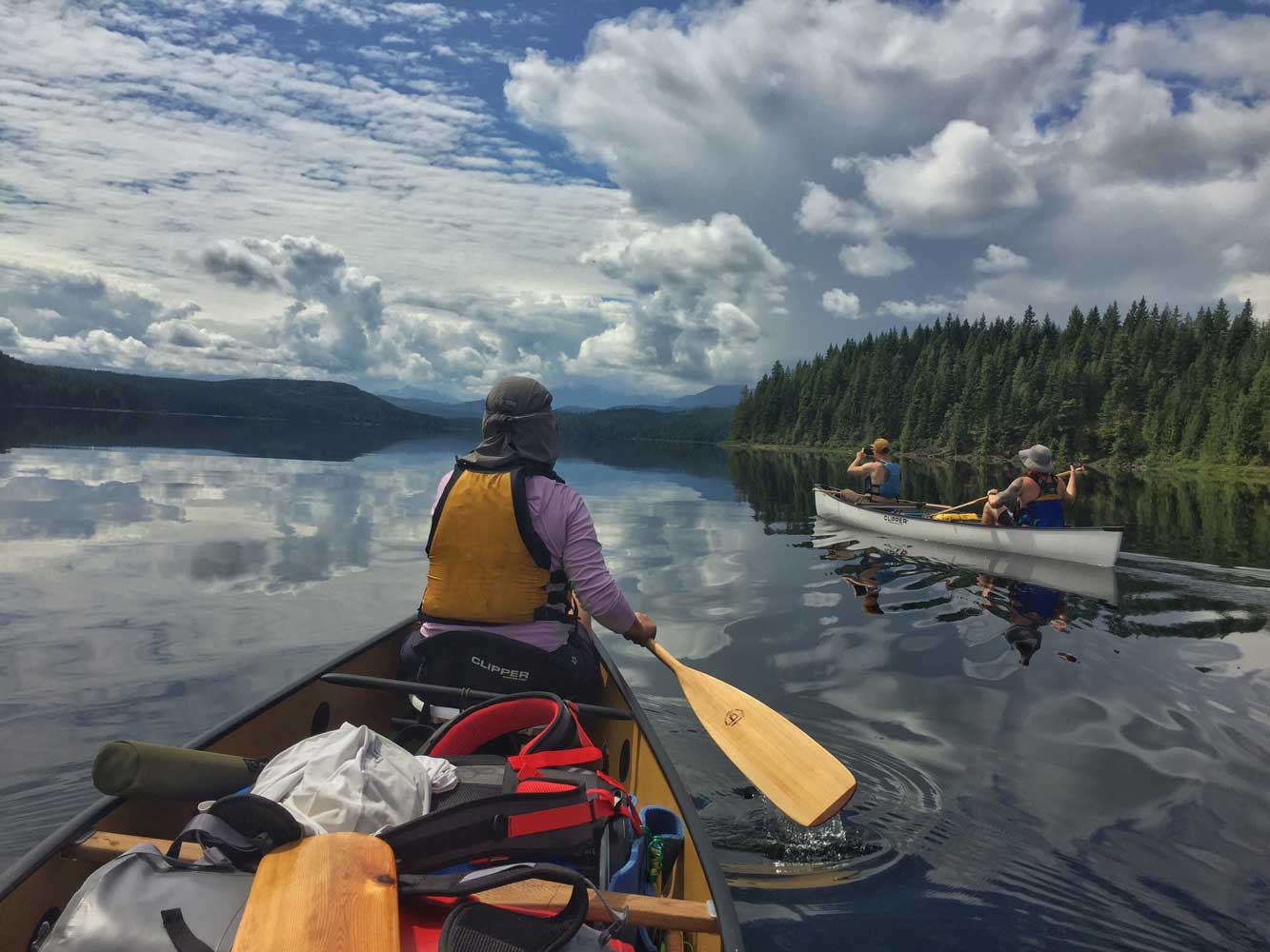 Kolme ihmistä melontaretkellä kahdella kanootilla kuusimetsien reunustamalla tyynellä järvellä kauniina kesäpäivänä.