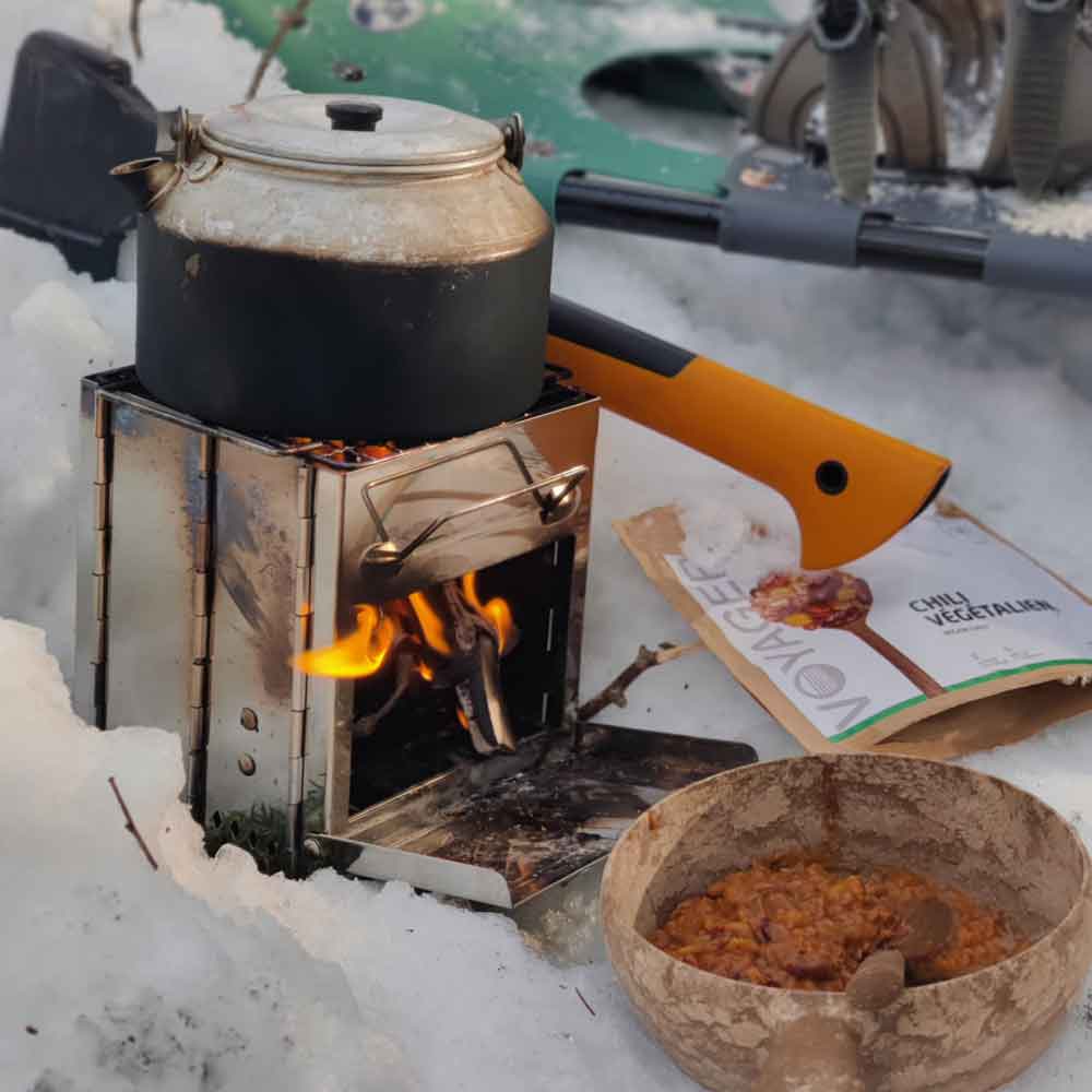 Chili sin carne retkiruoka kuplika-astissa risukeittimen edessä lumisessa maastossa. Taustalla retkiruokapussii, kirves ja lumikengät.