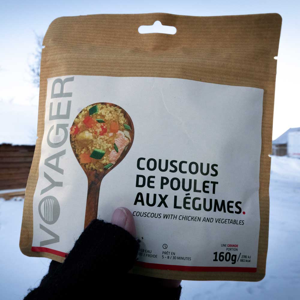 Kana-couscous ja vihanneksia retkiruokapussi lumisessa maisemassa kädessä, takana taloja.