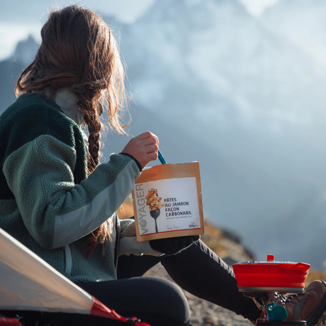Pitkälettinen nainen istuu teltan edessa ja syö retkiruoka Pasta carbonaraa kierratettävästä  retkiruokapussista.