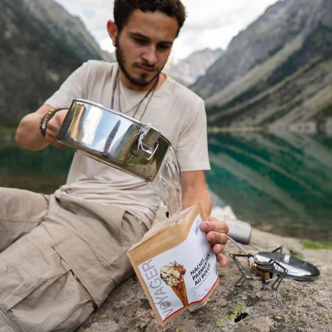 Mies vaaleassa asussa istuu vuoristossa järven vieressä kalliolla ja kaataa kattilasta vettä ranskalainen lisaperunasoselaatikko retkiruokaan kierrätettävässä pussissa.