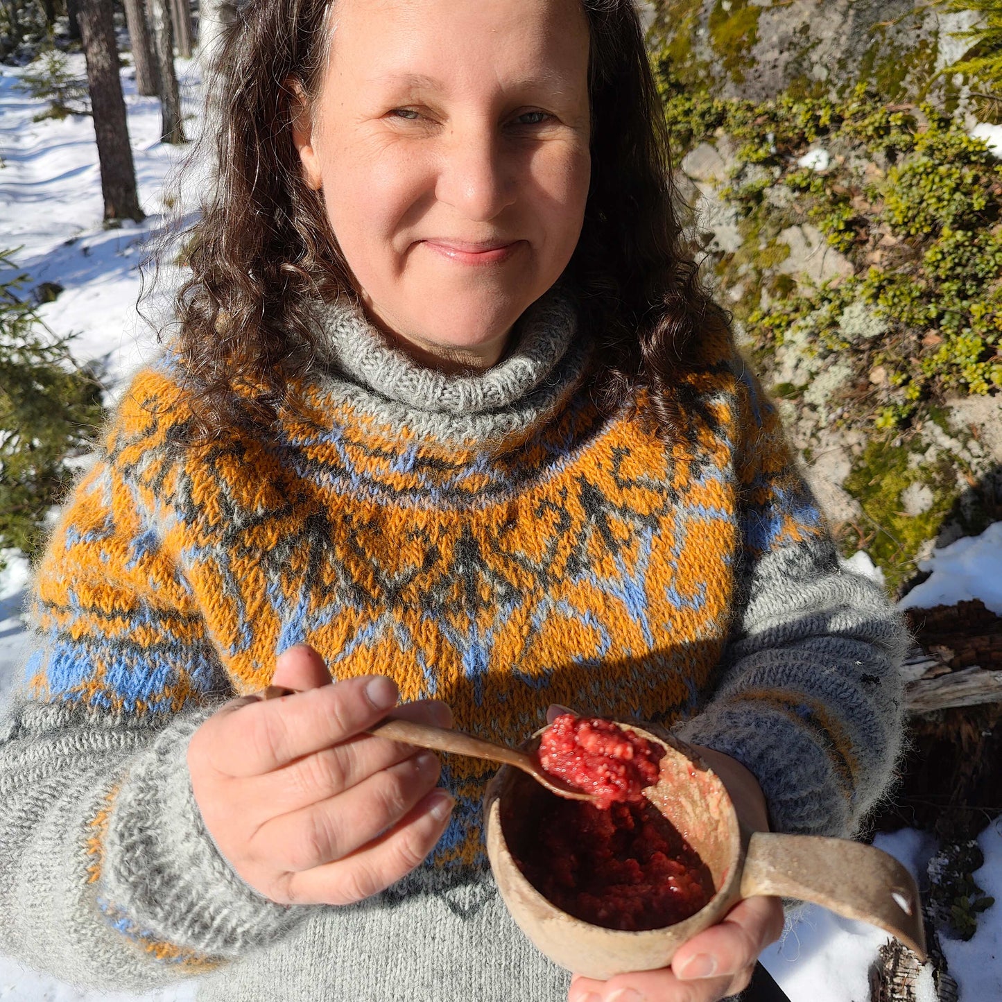 Omena-vadelma jälkiruokapaistos, retkiruoka valmiina kupilka-astiassa ja lusikassa tummatukkaisen harmaa villapaitaisen naisen kädessä lumisessa metsässä.