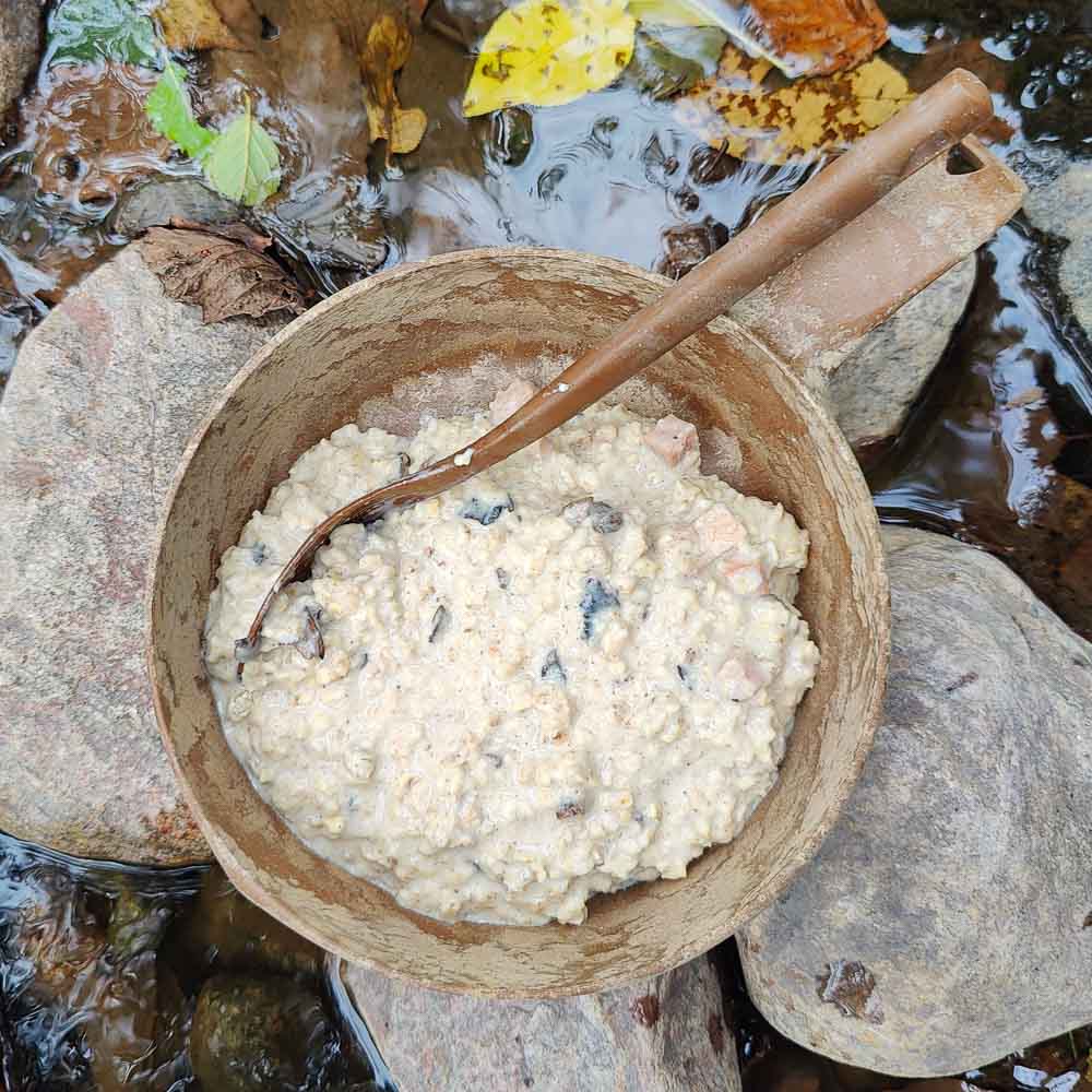 Vaeltajan savukinkku-herkkusienipuuro-retkiruoka valmiina Kuplikassa, jossa lusikka, kivien päällä veden ääressä.